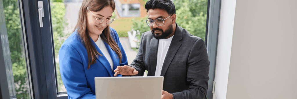 Two colleagues standing by a window, looking at a laptop together. They appear engaged in a collaborative discussion while reviewing information on the screen.