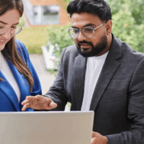 Two colleagues standing by a window, looking at a laptop together. They appear engaged in a collaborative discussion while reviewing information on the screen.