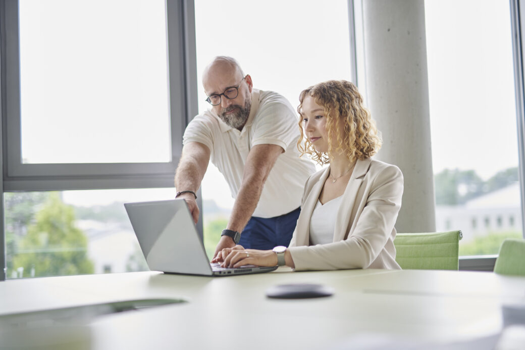 Two people are standing at a standing desk in front of a screen. In the background, two more people can be seen.